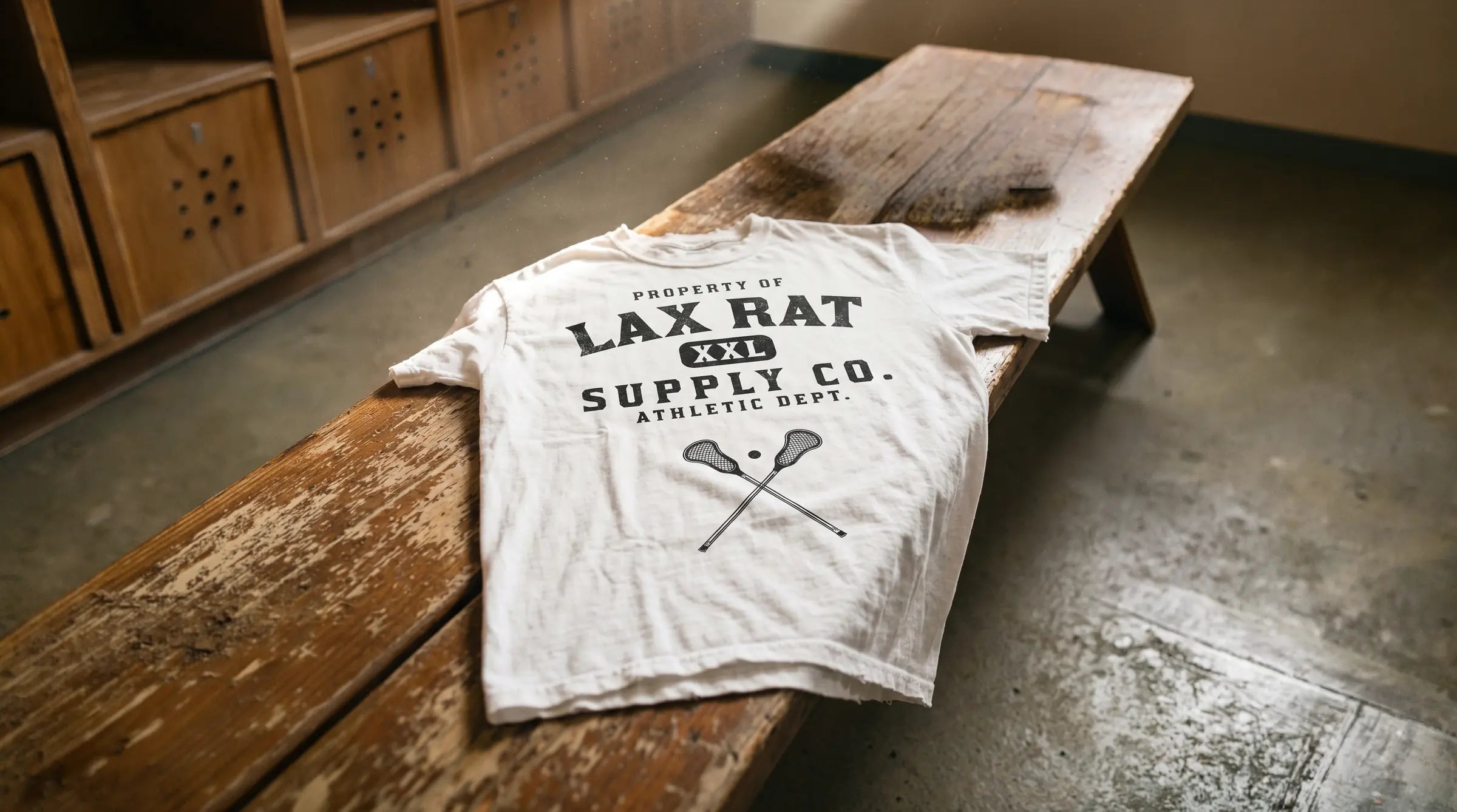 A white Lax Rat Supply Co. Athletic Dept. tee on a worn wooden locker room bench